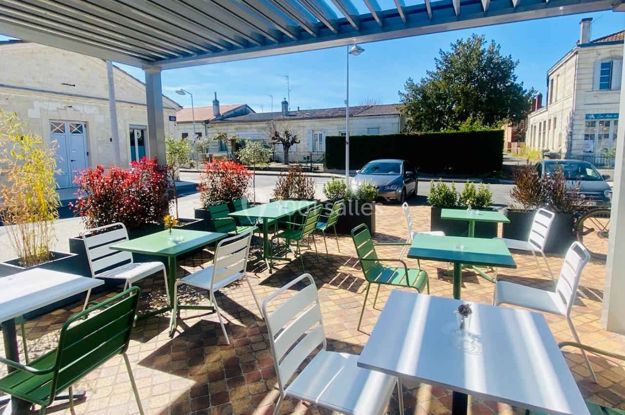 Terrasse extérieure avec tables et chaises colorées sous une pergola, entourée de plantes et donnant sur une rue calme.