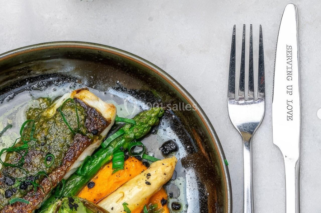 Assiette contenant du poisson grillé, asperges vertes et blanches, légumes et sauce, avec couteau et fourchette à côté.