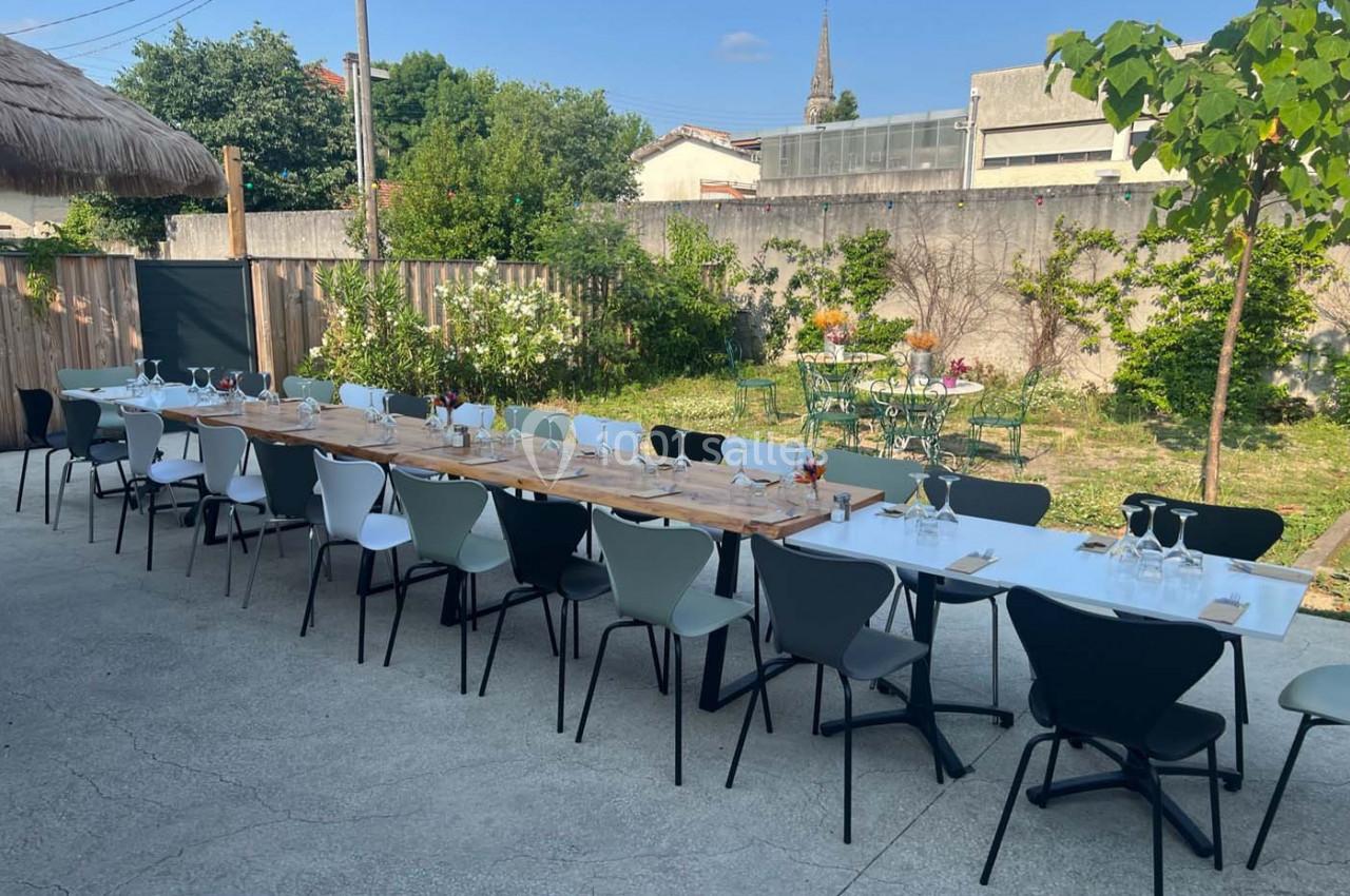 Tables dressées pour un repas en extérieur, alignées sur une terrasse avec un jardin et des arbres en arrière-plan.