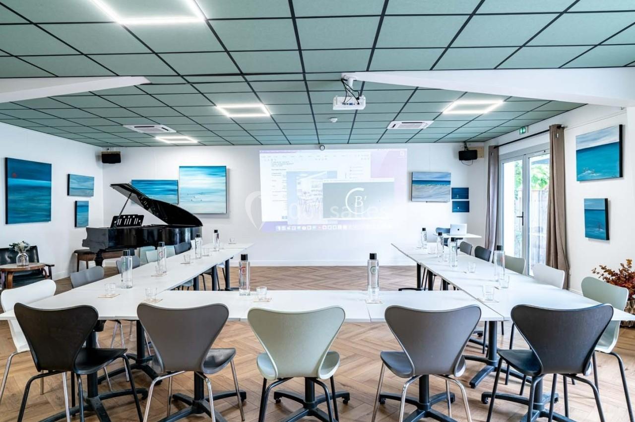 Salle de réunion lumineuse avec tables en U, chaises modernes, piano à queue et tableaux accrochés aux murs.