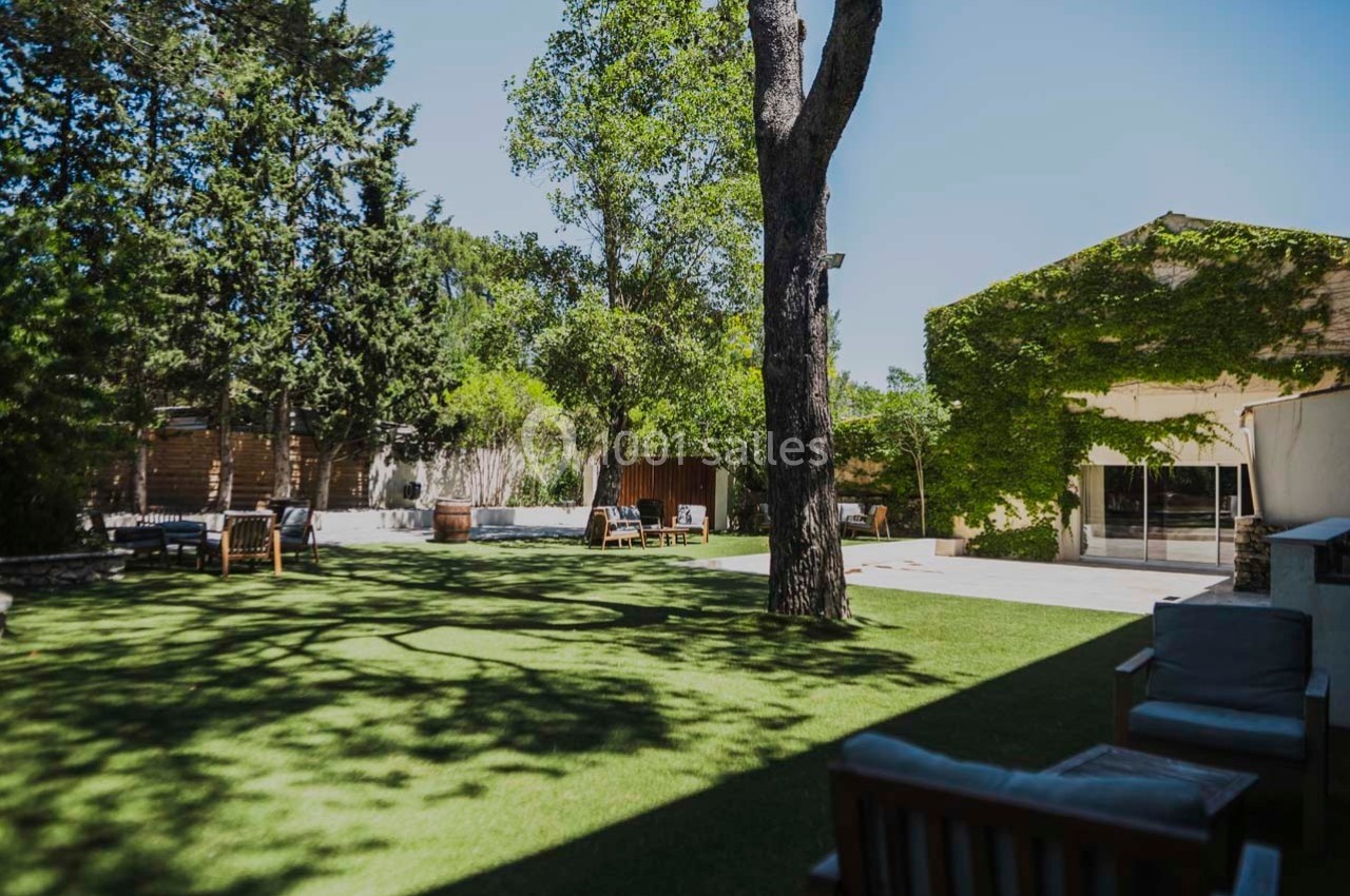 Jardin avec pelouse, arbres, mobilier en bois et bâtiment recouvert de végétation sous un ciel dégagé.