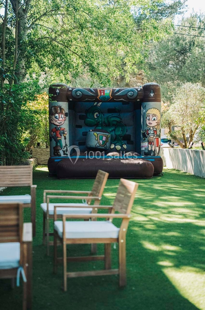 Château gonflable décoré sur le thème médiéval installé sur une pelouse, entouré d'arbres et de chaises en bois.