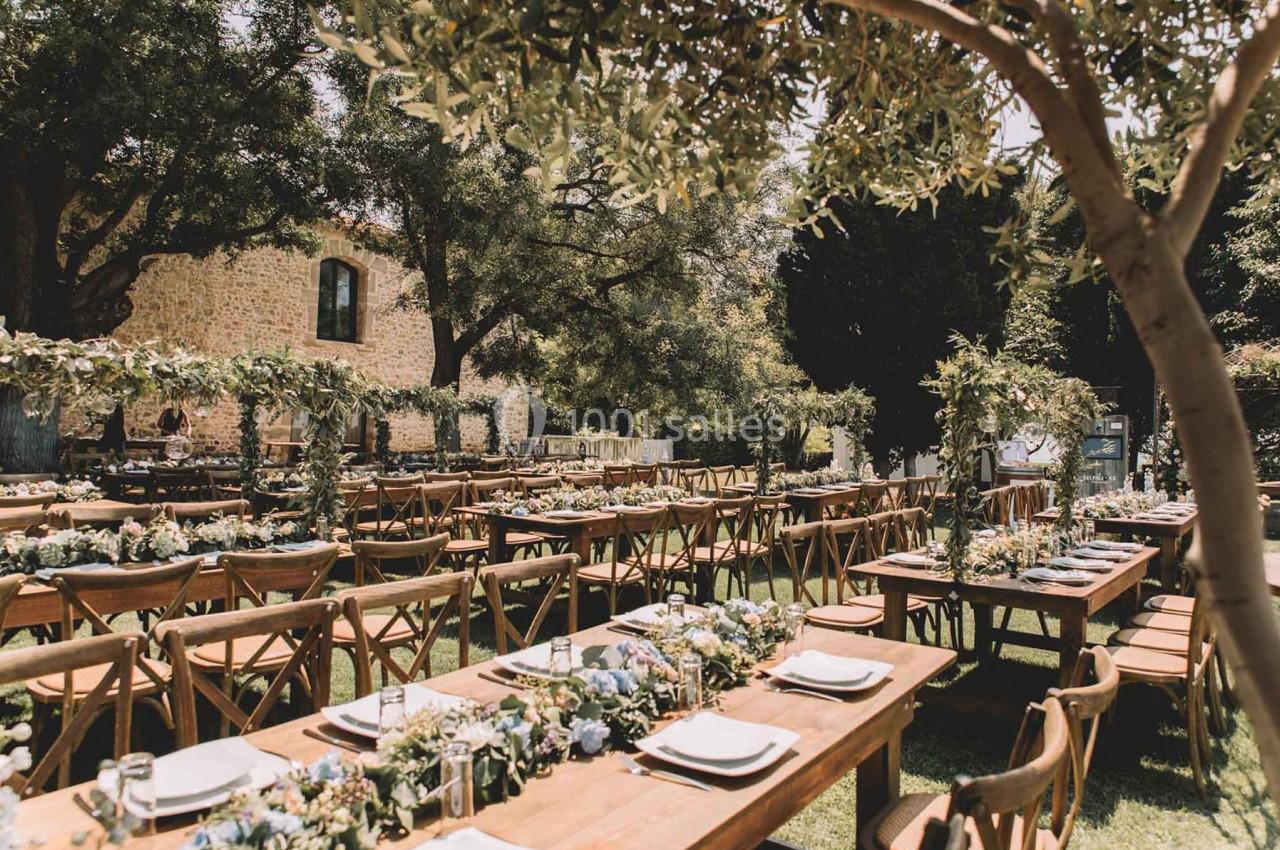 Tables en bois décorées de fleurs et de vaisselle, disposées en extérieur sous des arbres dans un cadre champêtre.