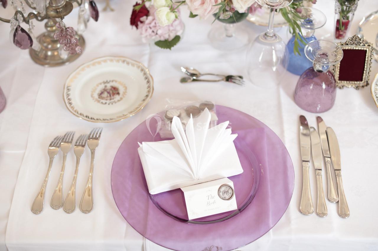 Table dressée avec assiette violette, serviette pliée, couverts en argent et décorations florales élégantes.