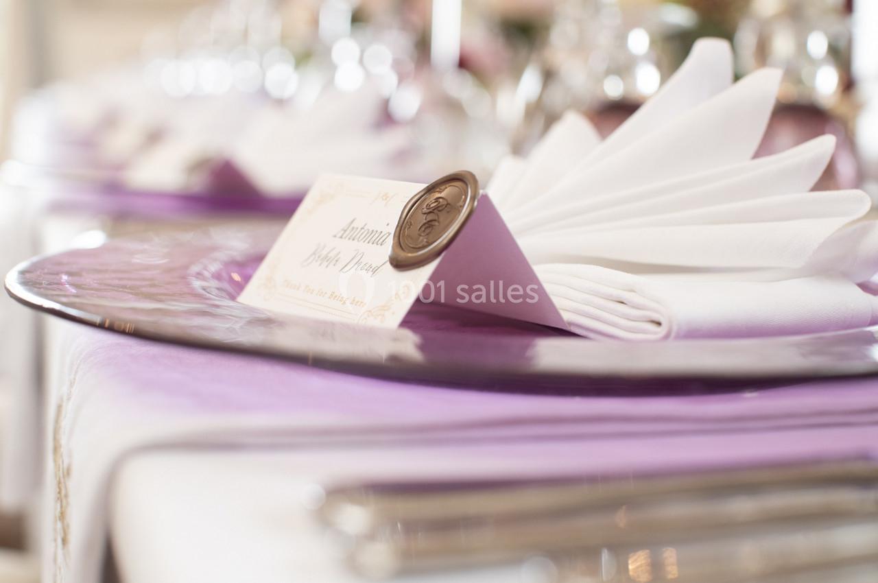 Serviette pliée avec une carte nominative et un sceau en cire, posée sur une assiette violette dans un décor de table…