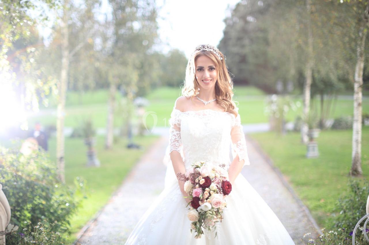 Femme en robe de mariée tenant un bouquet de fleurs, debout sur une allée bordée d'arbres dans un jardin lumineux.