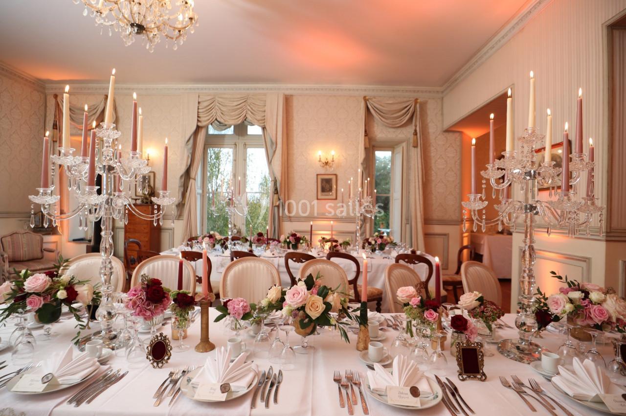 Salle élégante décorée pour un dîner, avec chandeliers, fleurs fraîches et tables dressées avec vaisselle raffinée.