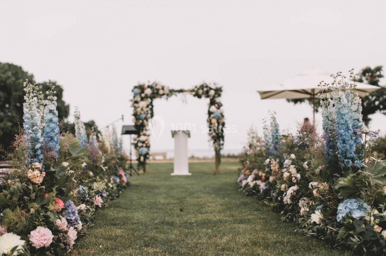 Allée bordée de fleurs menant à une arche décorée pour une cérémonie en extérieur sur pelouse.
