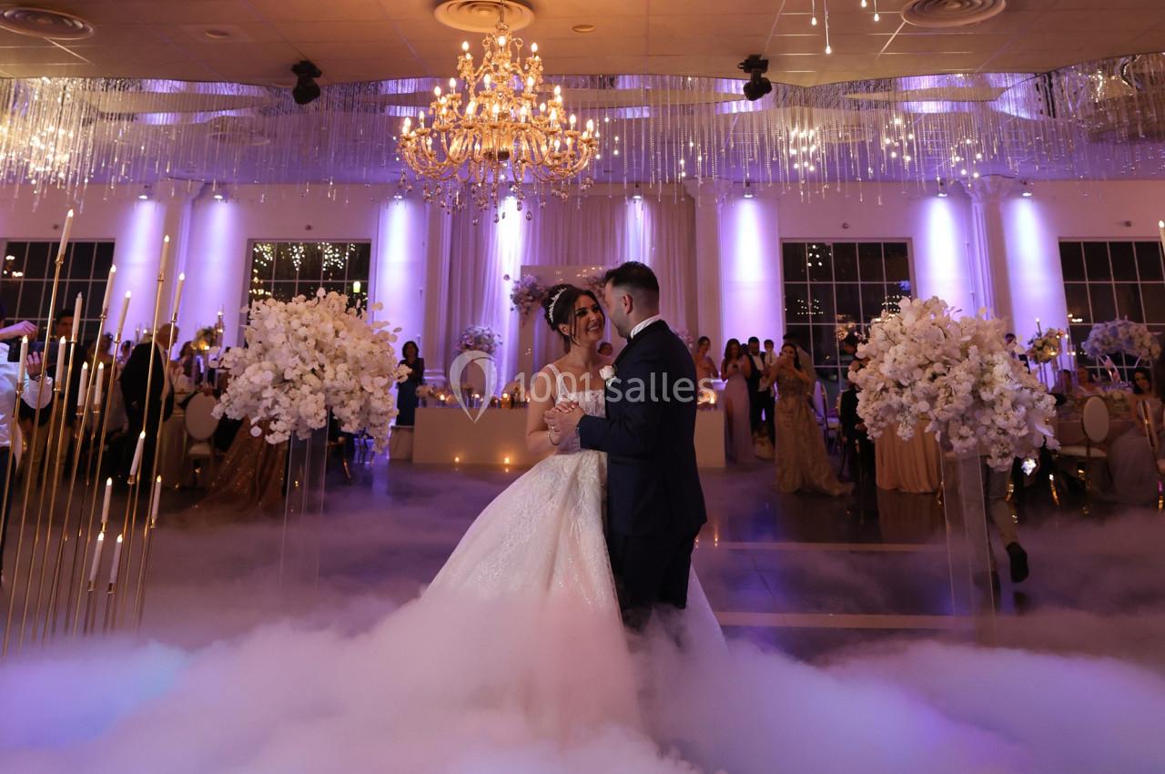 Un couple danse leur première danse de mariage dans une salle élégante éclairée de lumières violettes et dorées.