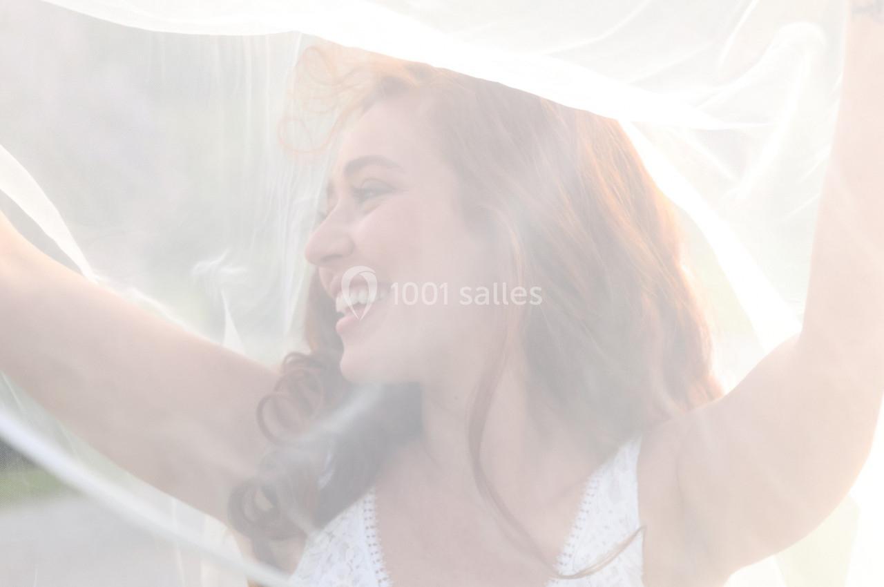 Femme souriante en robe blanche, partiellement voilée par un tissu translucide dans une lumière douce.