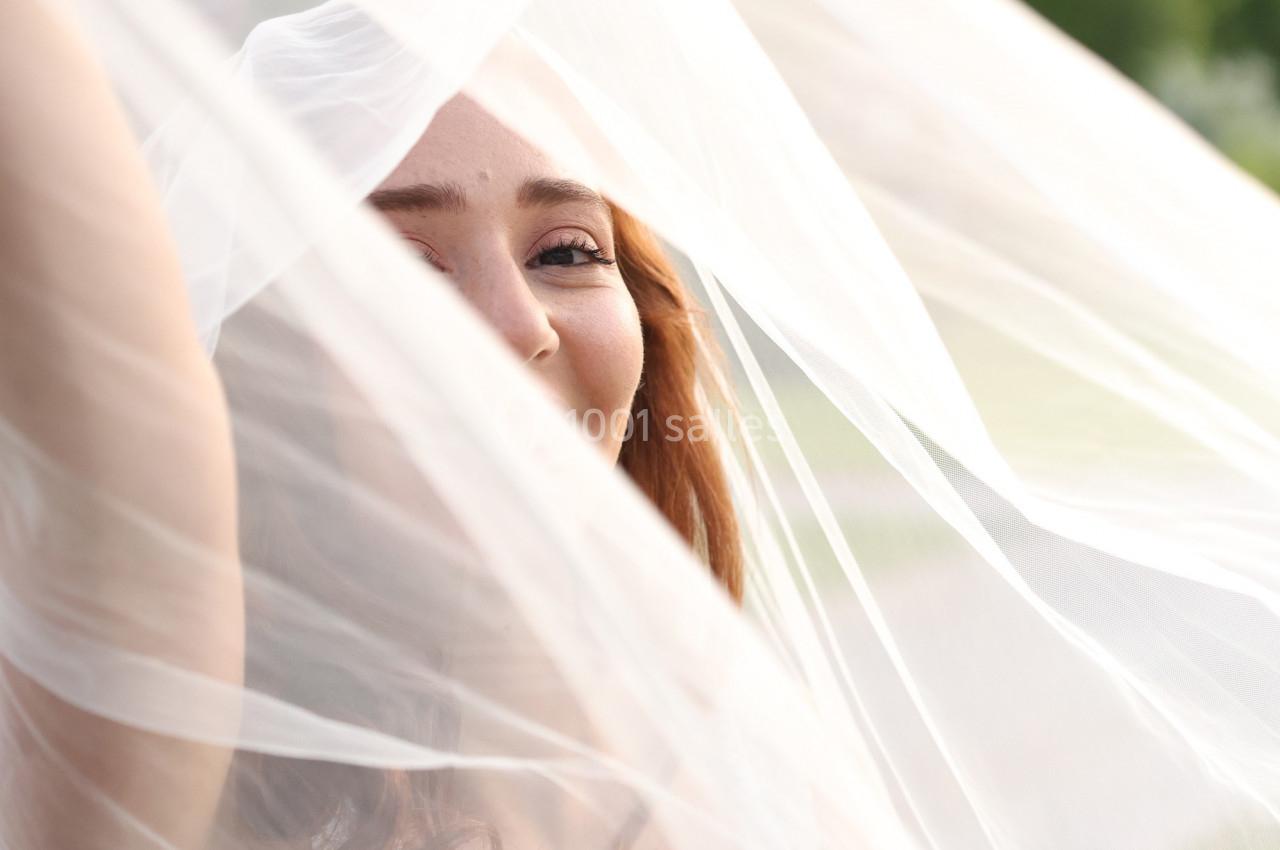 Femme souriante partiellement cachée derrière un voile blanc translucide, en extérieur lumineux.