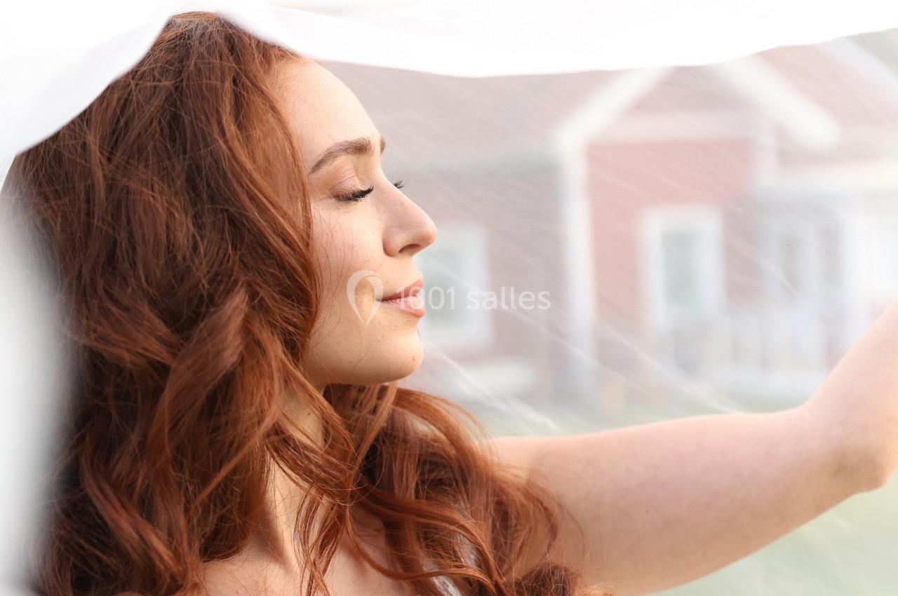 Femme aux cheveux roux souriant légèrement, vue de profil, sous un voile blanc avec des maisons floues en arrière-plan.