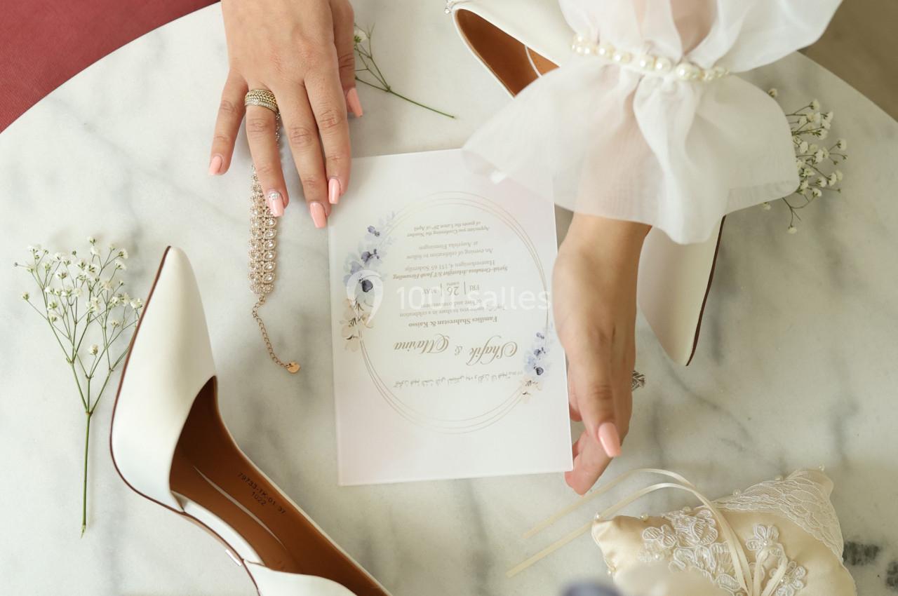 Mains ajustant une invitation de mariage sur une table en marbre, entourée de chaussures, bijoux et fleurs délicates.