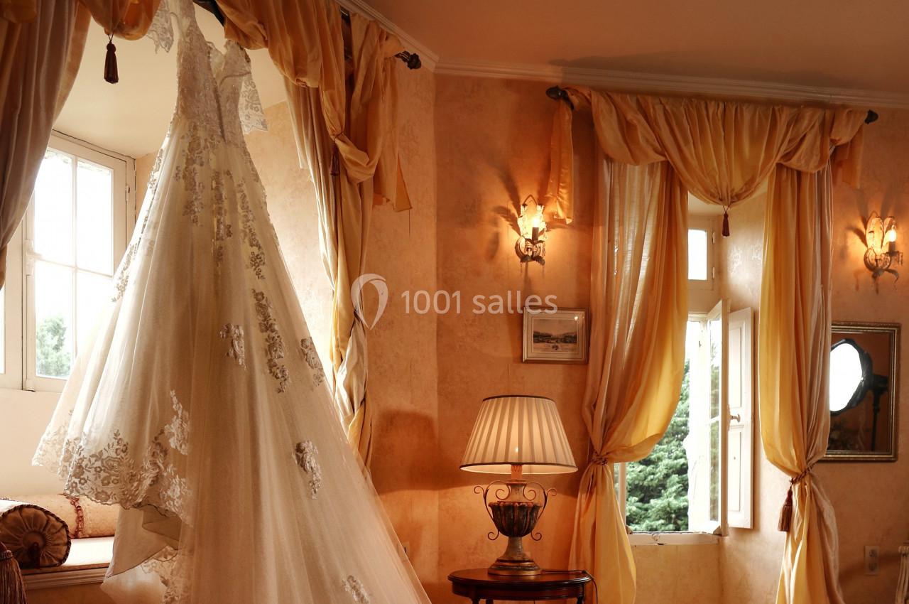 Robe de mariée en dentelle suspendue près d'une fenêtre dans une pièce lumineuse aux rideaux dorés.