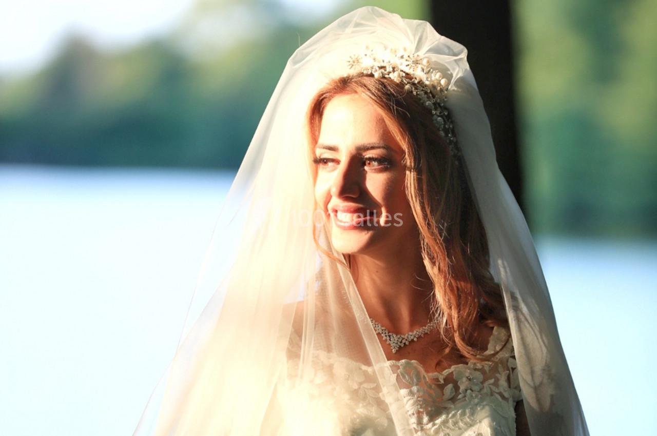 Une femme en robe de mariée et voile sourit, éclairée par une lumière naturelle près d'un plan d'eau.