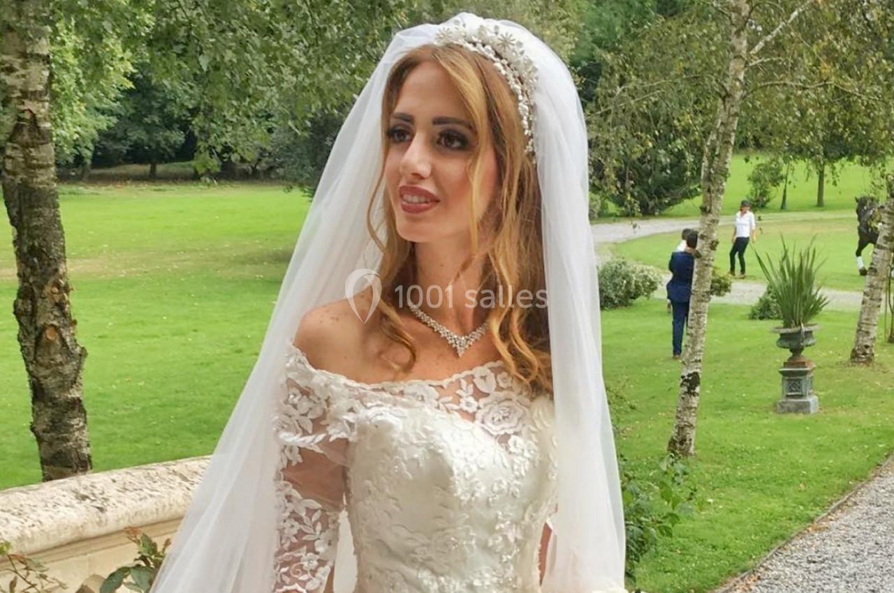 Une mariée en robe blanche et voile se tient dans un jardin verdoyant, entourée d'arbres et d'allées.