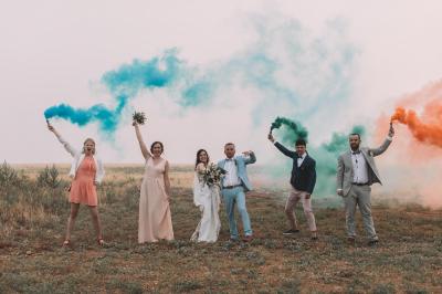 Un groupe de personnes en tenue de cérémonie tient des fumigènes colorés dans un champ ouvert.