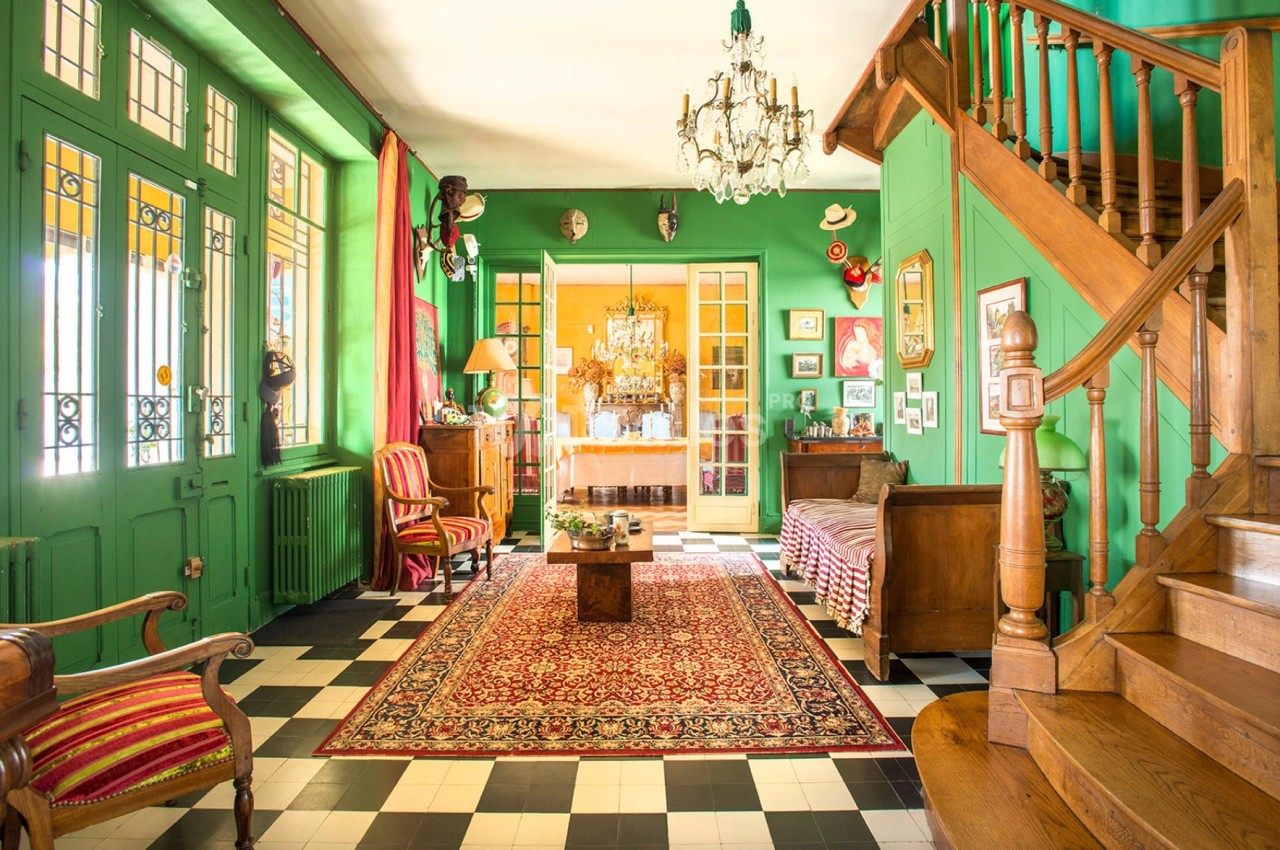 Salon lumineux avec murs verts, escalier en bois, tapis rouge, meubles anciens et portes vitrées donnant sur une autre pièce.