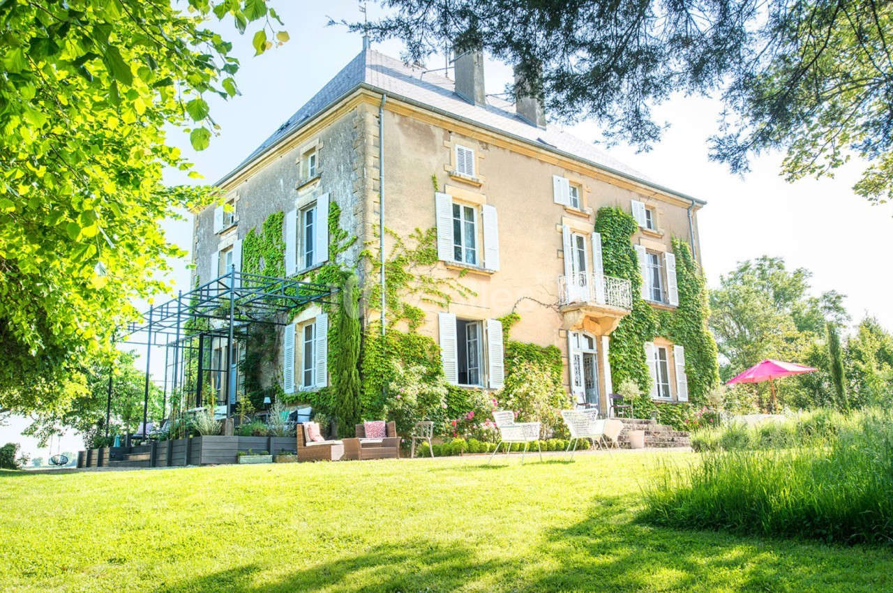 Grande maison en pierre avec volets blancs, entourée de verdure et d'un jardin ensoleillé avec mobilier extérieur.