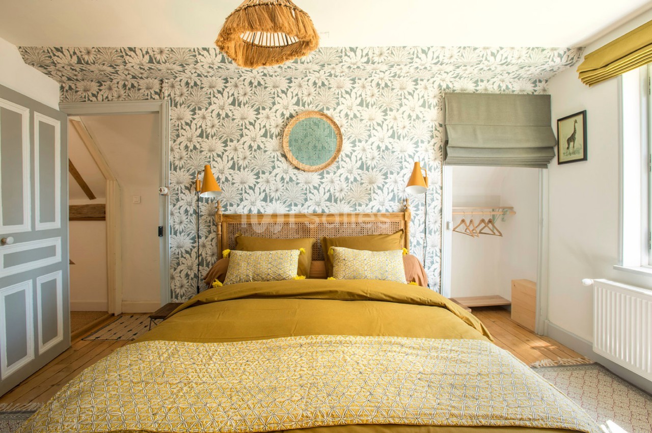 Chambre lumineuse avec lit double, linge de lit jaune, papier peint à motifs végétaux et décoration en bois naturel.