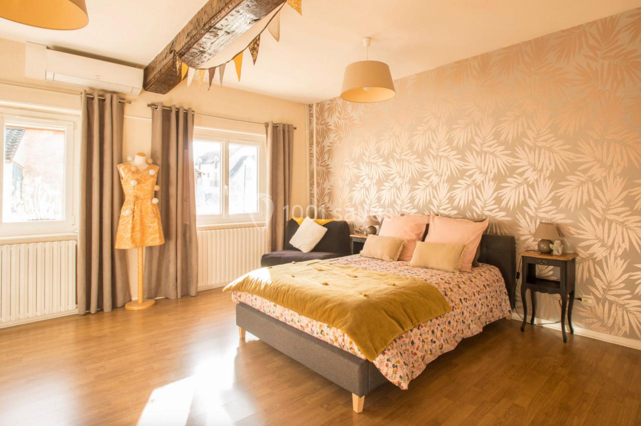 Chambre lumineuse avec lit double, parquet, mur décoré de motifs végétaux, mannequin et grandes fenêtres avec rideaux.