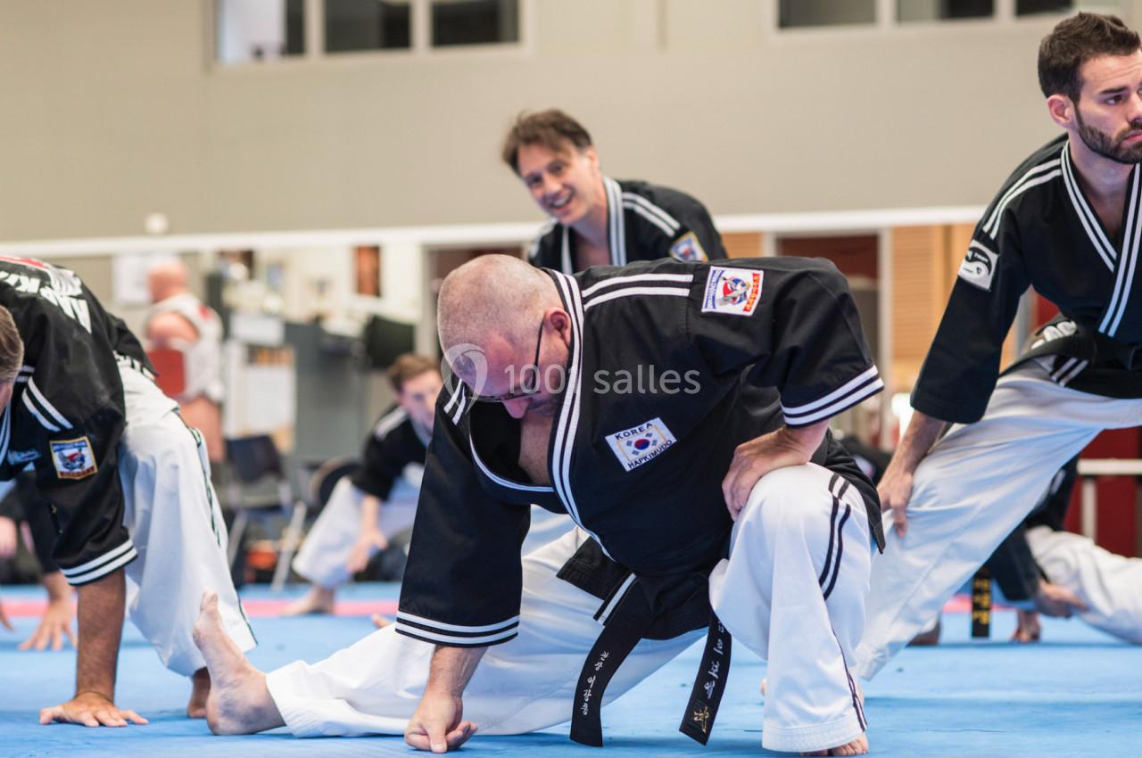 Des personnes en tenue d'arts martiaux effectuent des étirements sur un sol bleu dans une salle d'entraînement.