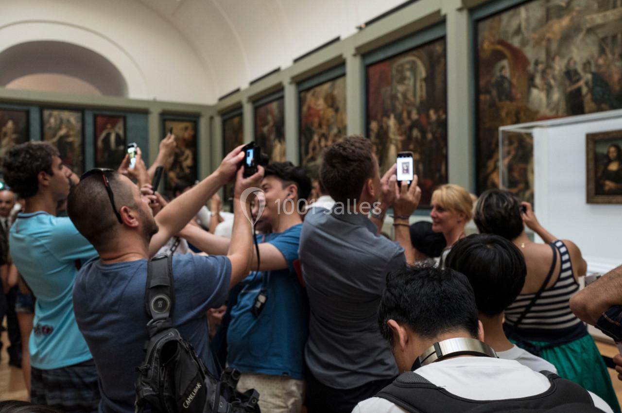 Des visiteurs photographient une œuvre d'art dans une galerie bondée, entourés de peintures exposées sur les murs.