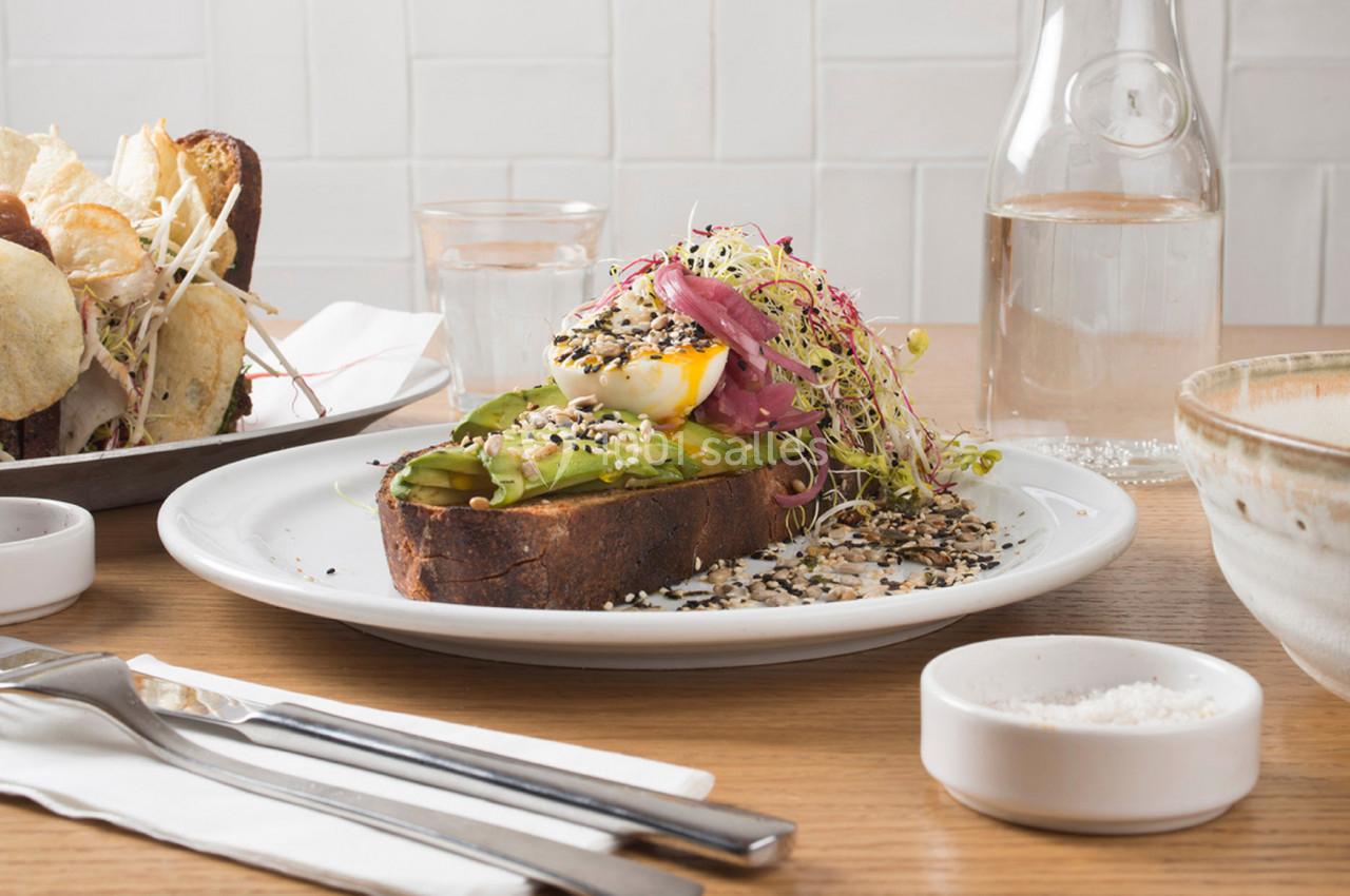 Une tartine garnie d'avocat, œuf poché, graines et pousses, servie sur une table avec d'autres plats et couverts.