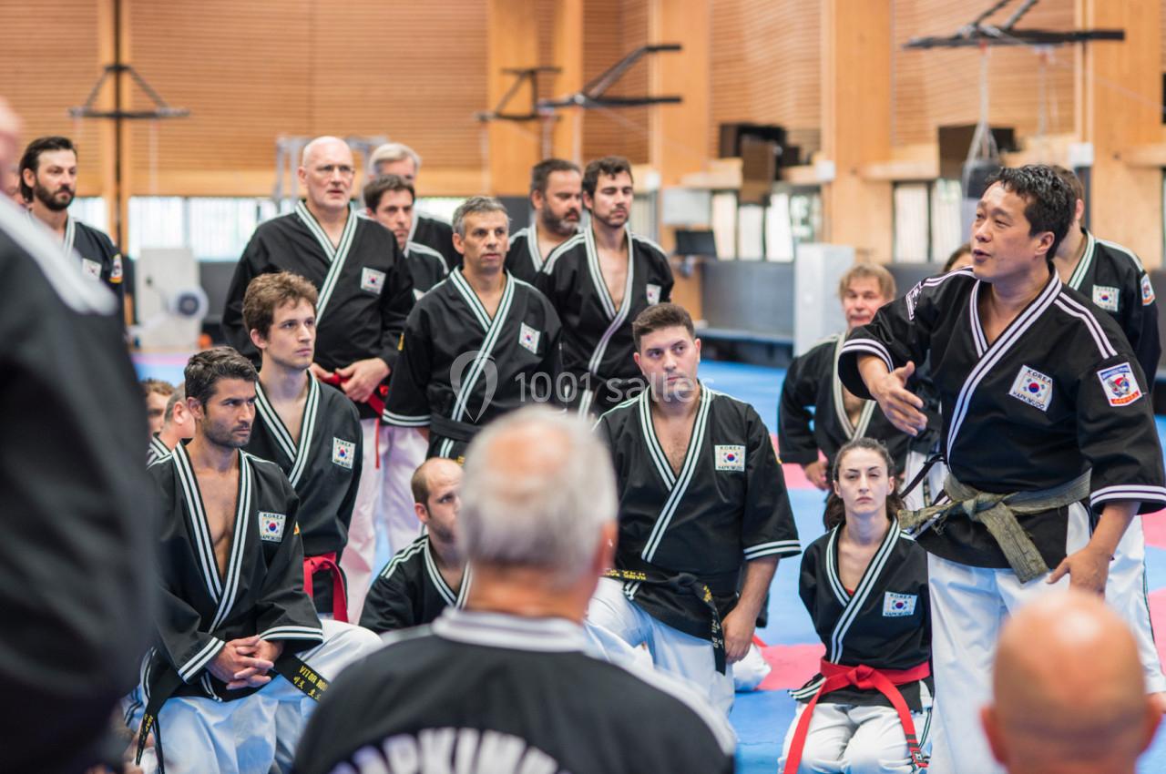 Un groupe de pratiquants d'arts martiaux en uniforme écoute attentivement un instructeur dans un gymnase.