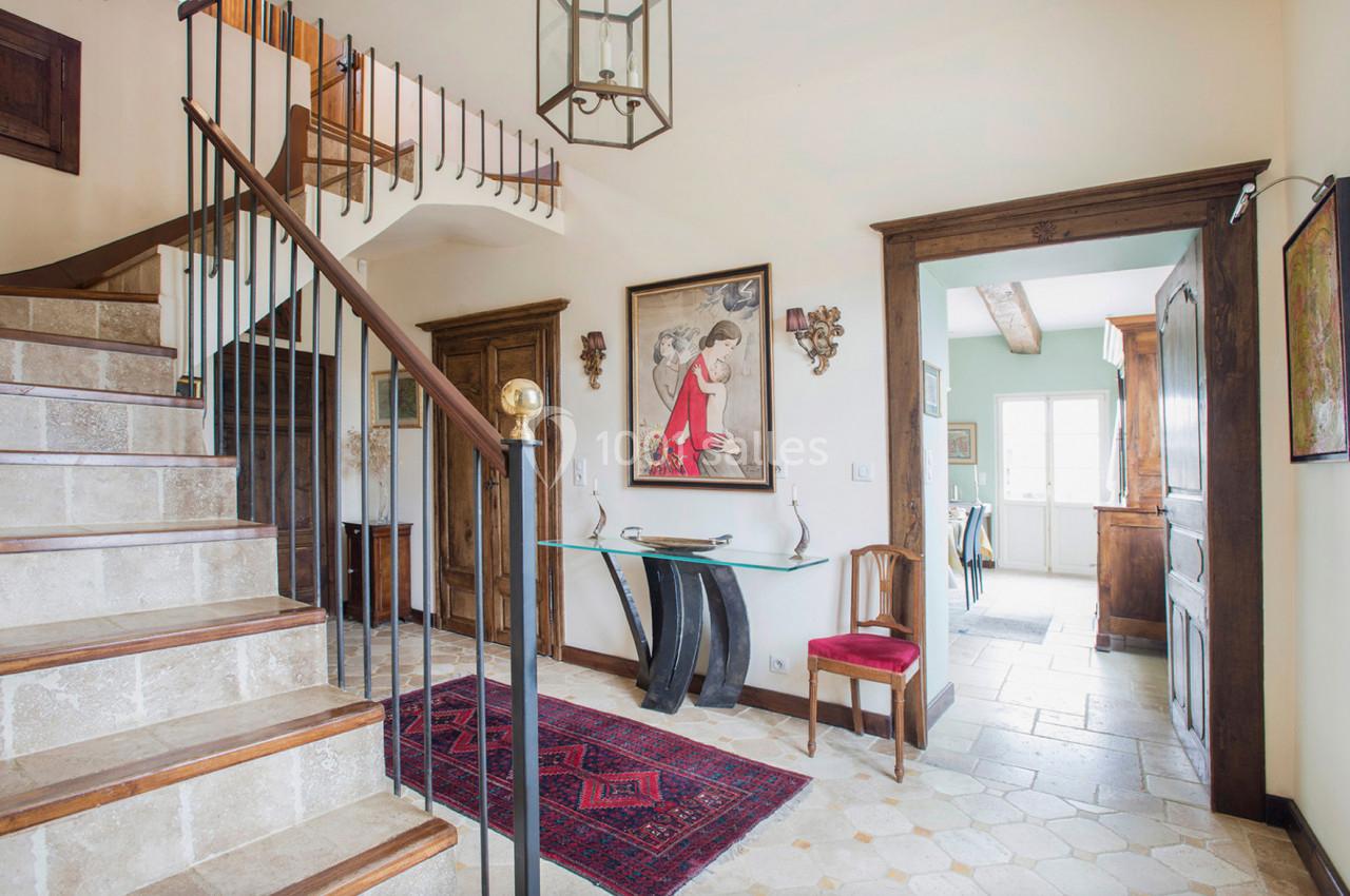 Entrée lumineuse avec escalier en pierre, tapis rouge, meubles en bois et accès à une cuisine au fond.