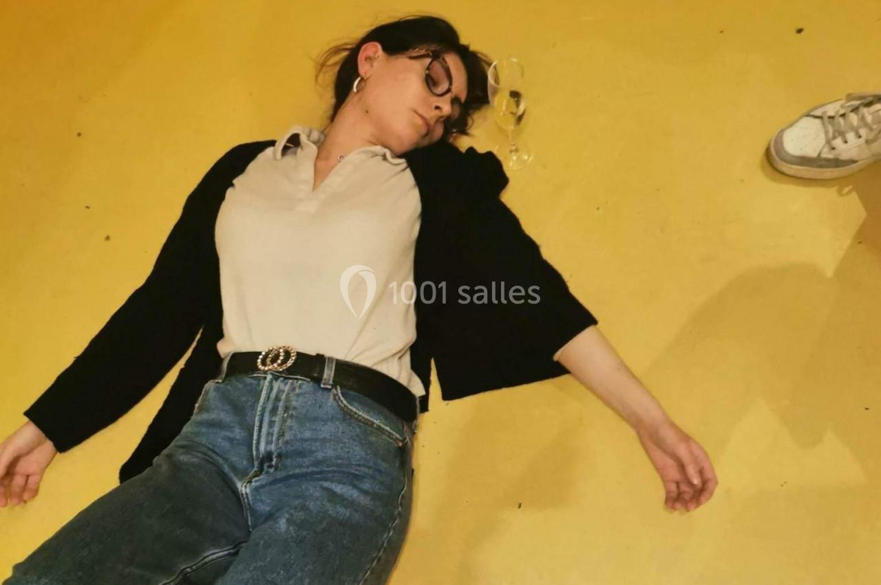 Une femme allongée sur le sol jaune, les lunettes tombées à côté d'elle, avec une chaussure visible en arrière-plan.