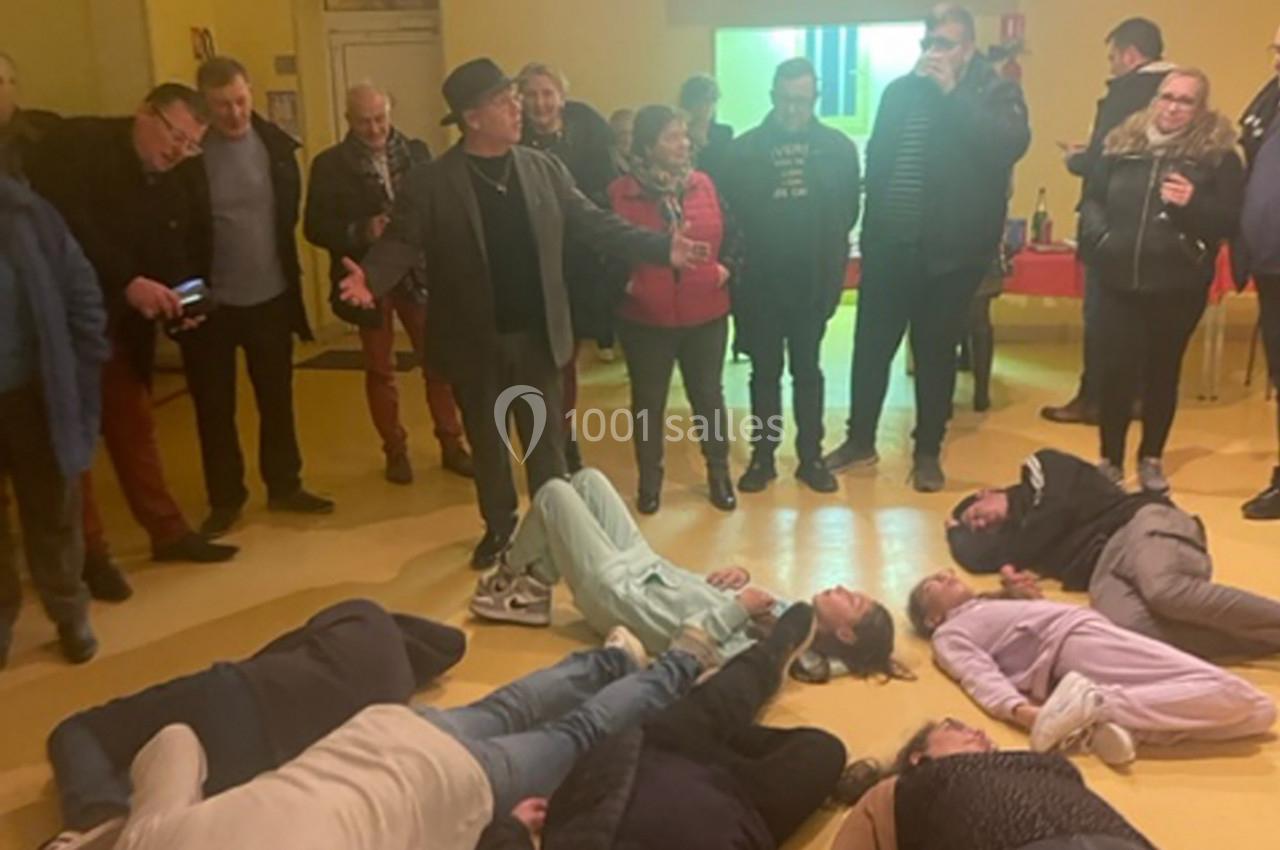 Un groupe de personnes debout observe plusieurs individus allongés sur le sol dans une salle éclairée.