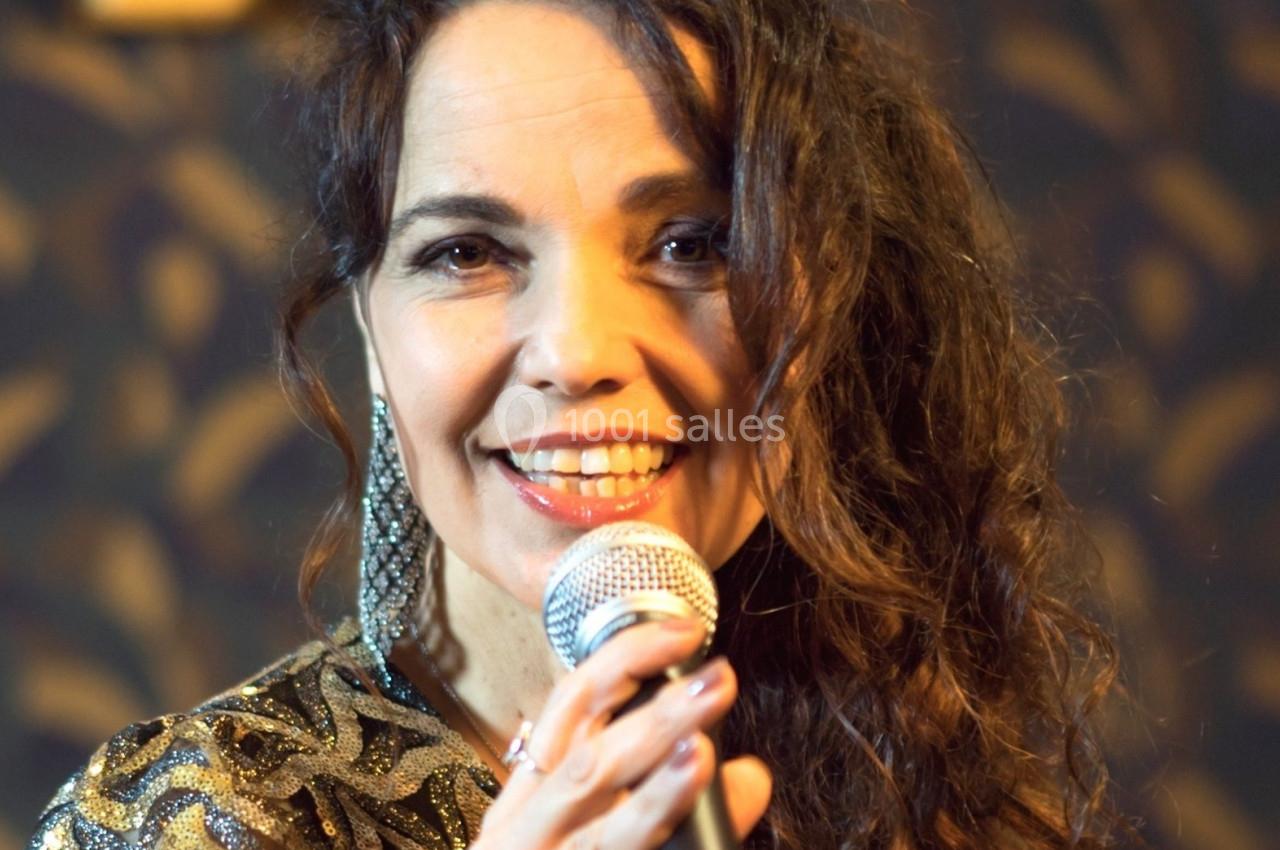 Femme souriante tenant un micro, vêtue d'une robe à motifs dorés, devant un fond sombre à motifs.