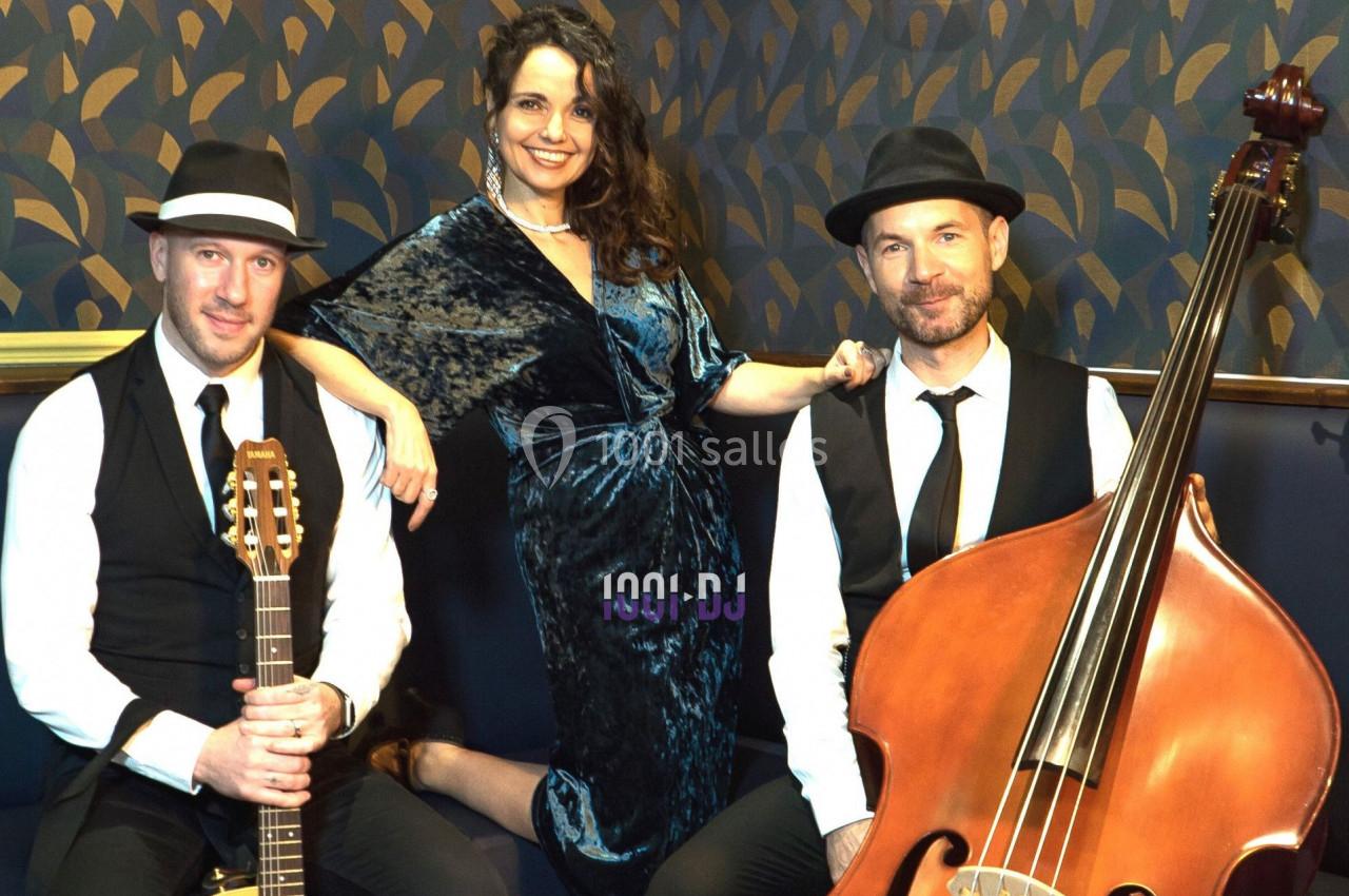 Un trio musical pose avec une guitare, une contrebasse et une chanteuse en robe bleue devant un mur décoratif.