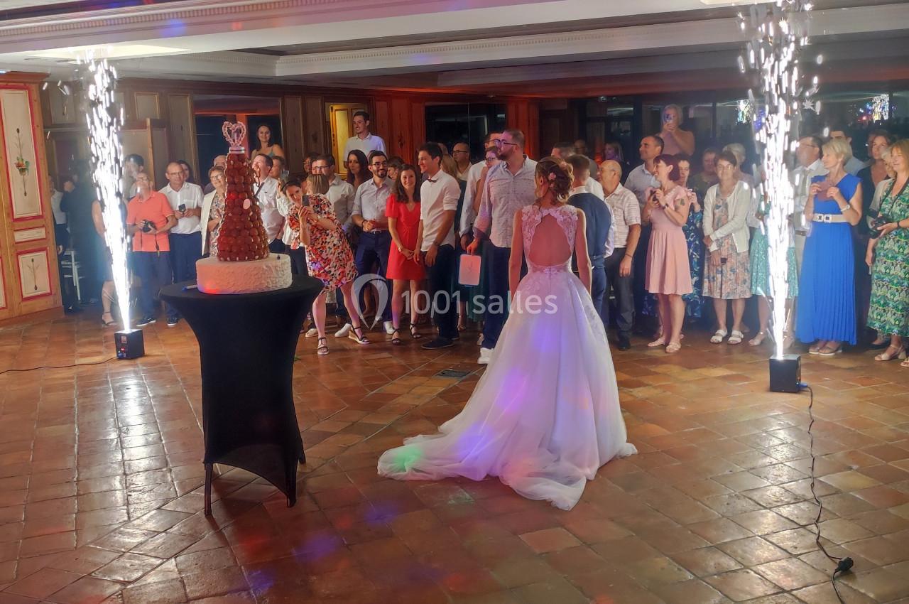Une mariée en robe blanche regarde un gâteau de mariage entouré d'invités dans une salle décorée avec des étincelles.