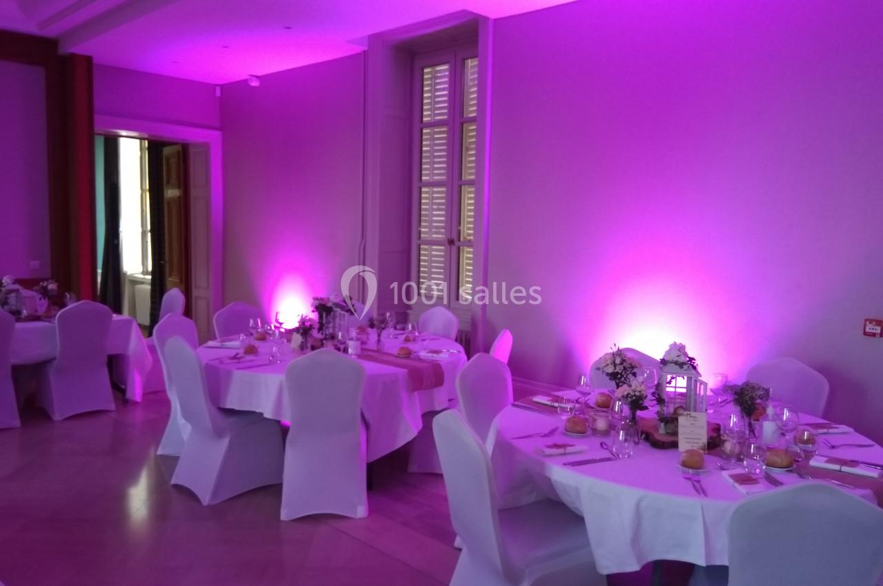 Salle de réception avec tables rondes dressées, nappes blanches et éclairage d'ambiance rose.