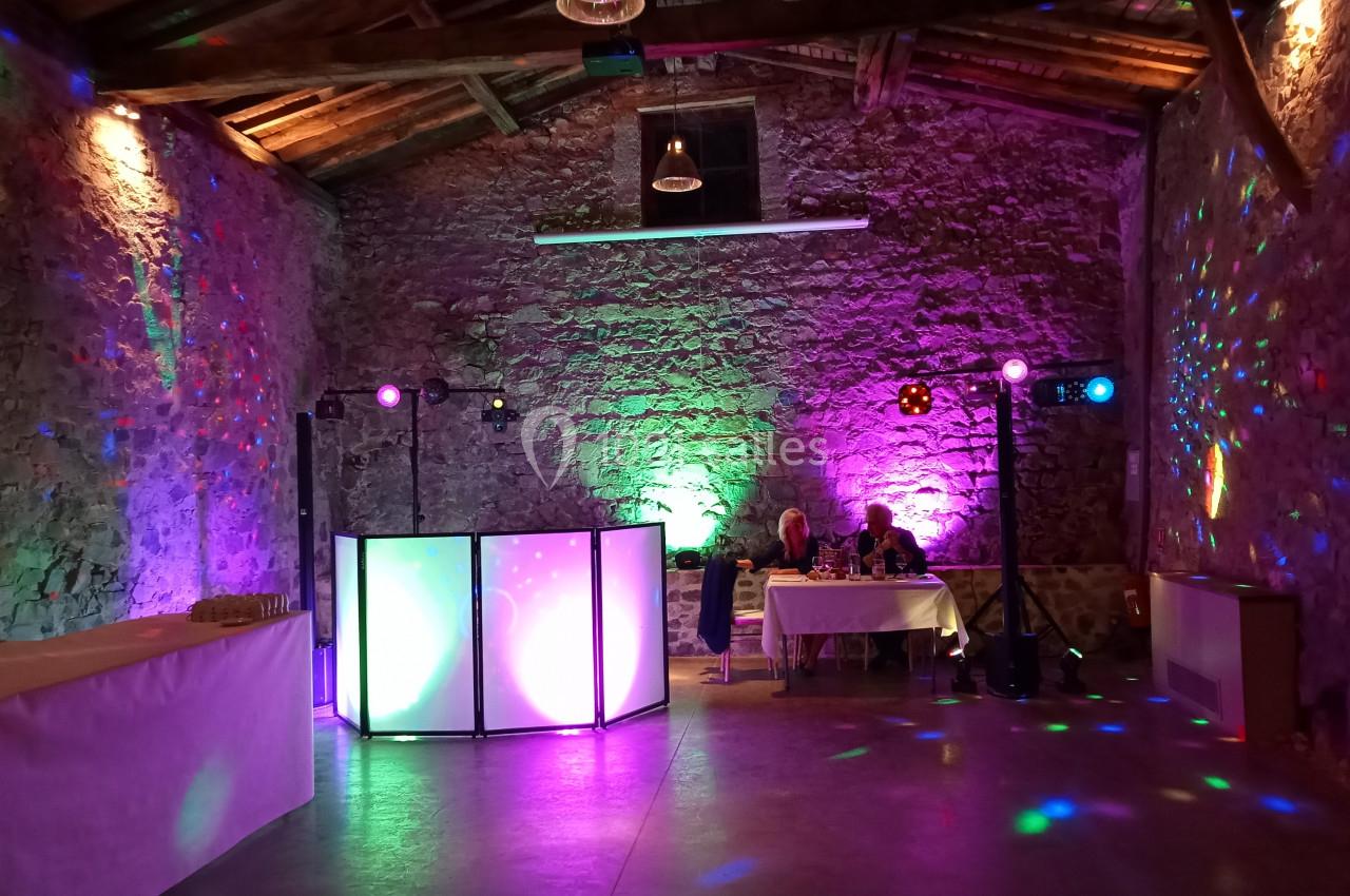 Salle en pierre éclairée par des lumières colorées, avec une table de mixage et une table dressée au fond.