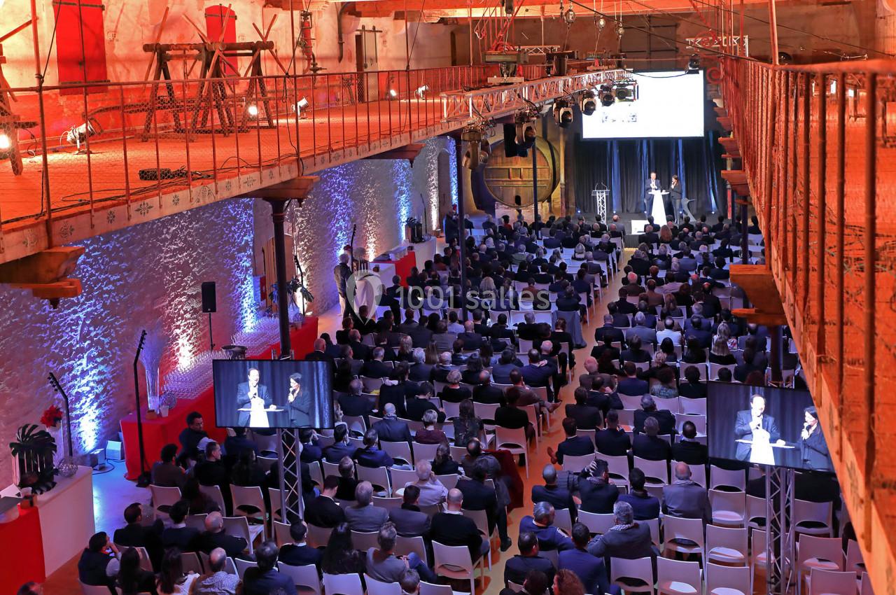 Salle comble avec des participants assis, regardant une présentation sur scène, éclairée par des projecteurs.
