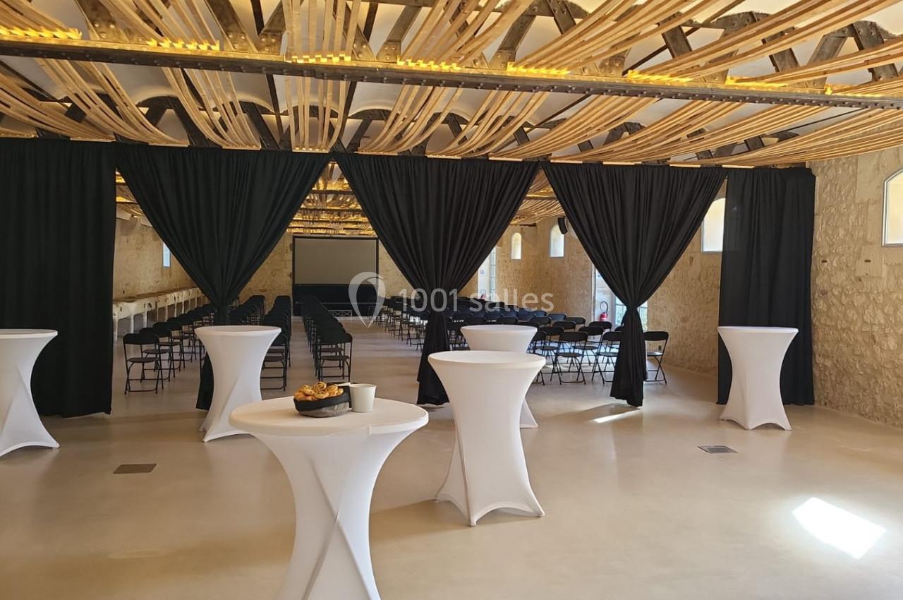 Salle en pierre avec des tables hautes drapées de blanc, rideaux noirs ouverts et rangées de chaises devant un écran.