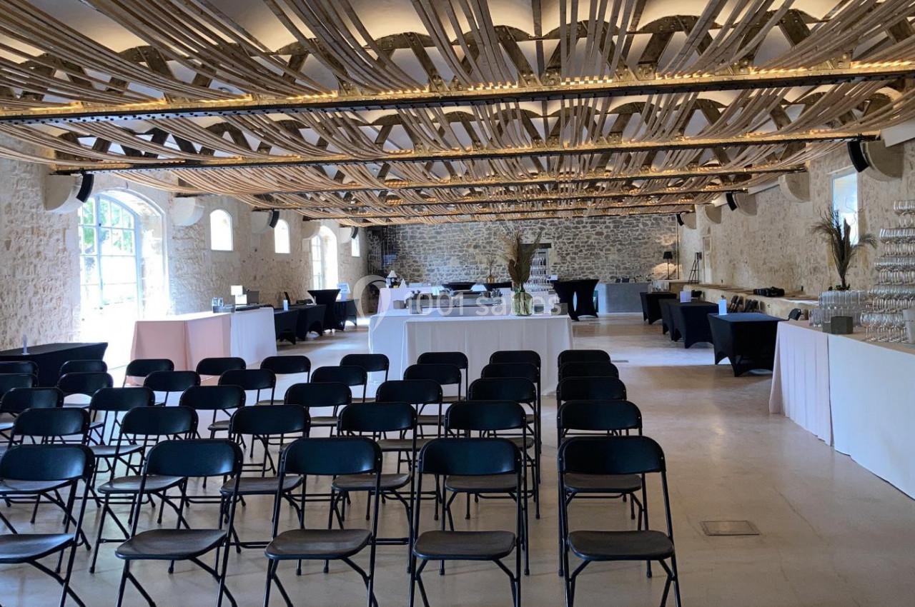 Salle en pierre avec des chaises noires alignées face à des tables dressées, éclairée par des fenêtres et un plafond…