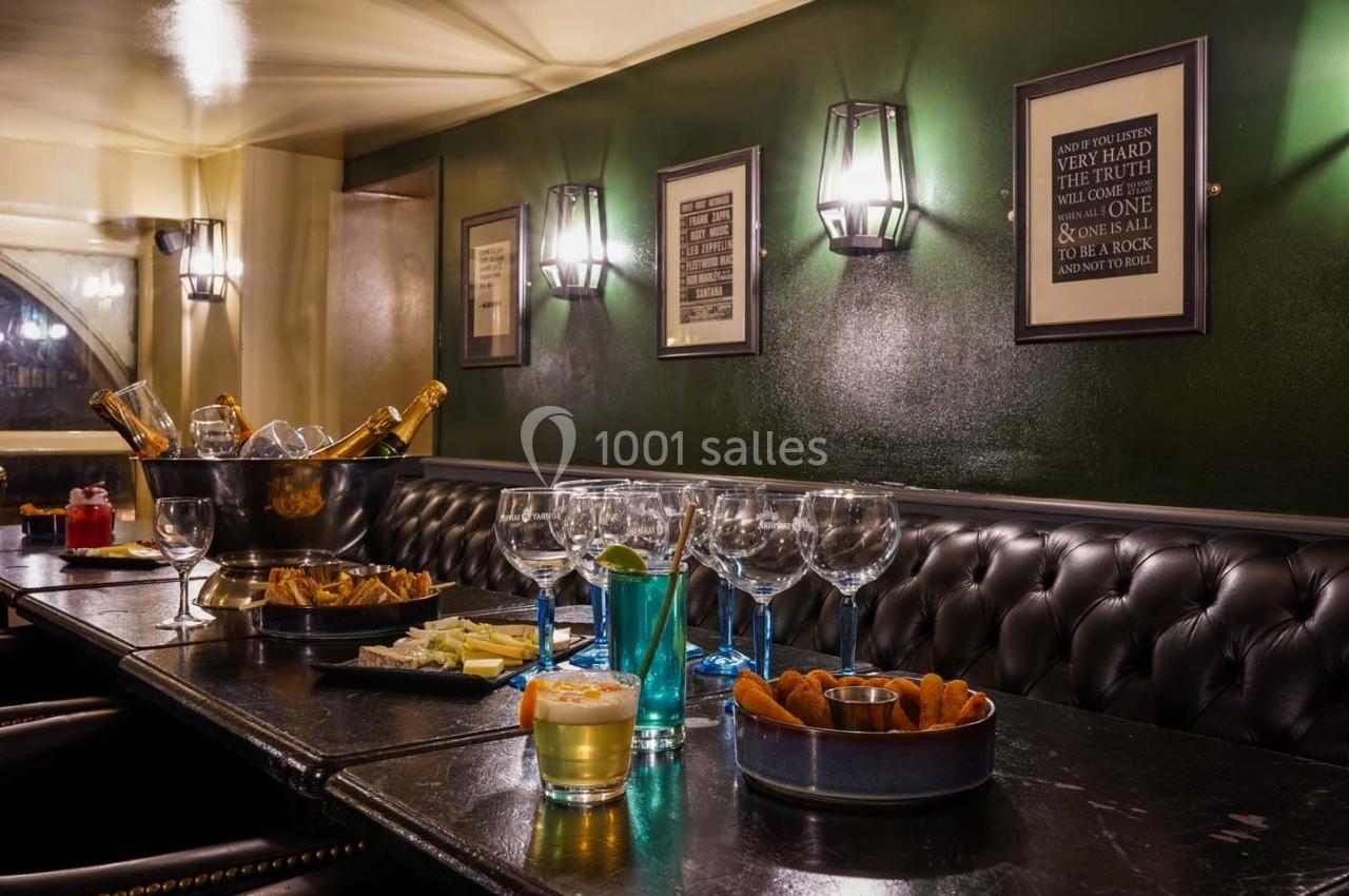 Salle avec banquettes en cuir, table dressée avec verres, bouteilles, amuse-bouches et éclairage mural tamisé.