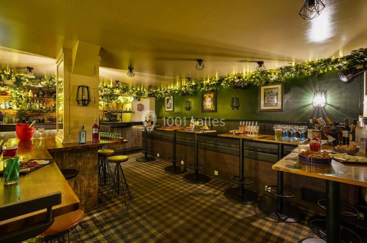Salle de bar chaleureuse avec tables hautes, banquettes, décorations végétales et éclairage tamisé.