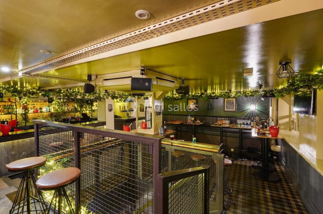 Intérieur d'un bar avec des tables hautes, un comptoir éclairé et une décoration végétale suspendue.