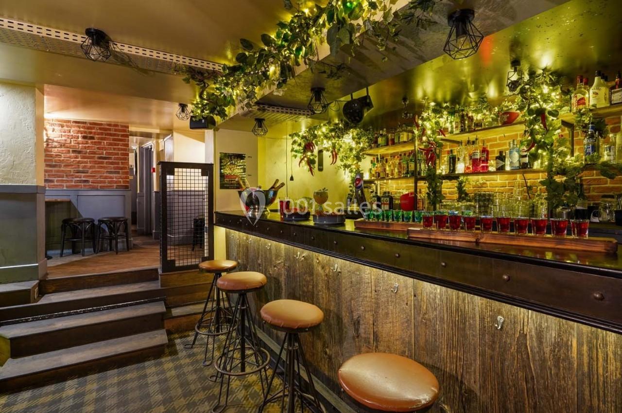 Bar chaleureux avec comptoir en bois, tabourets hauts, étagères garnies de bouteilles et décorations lumineuses.