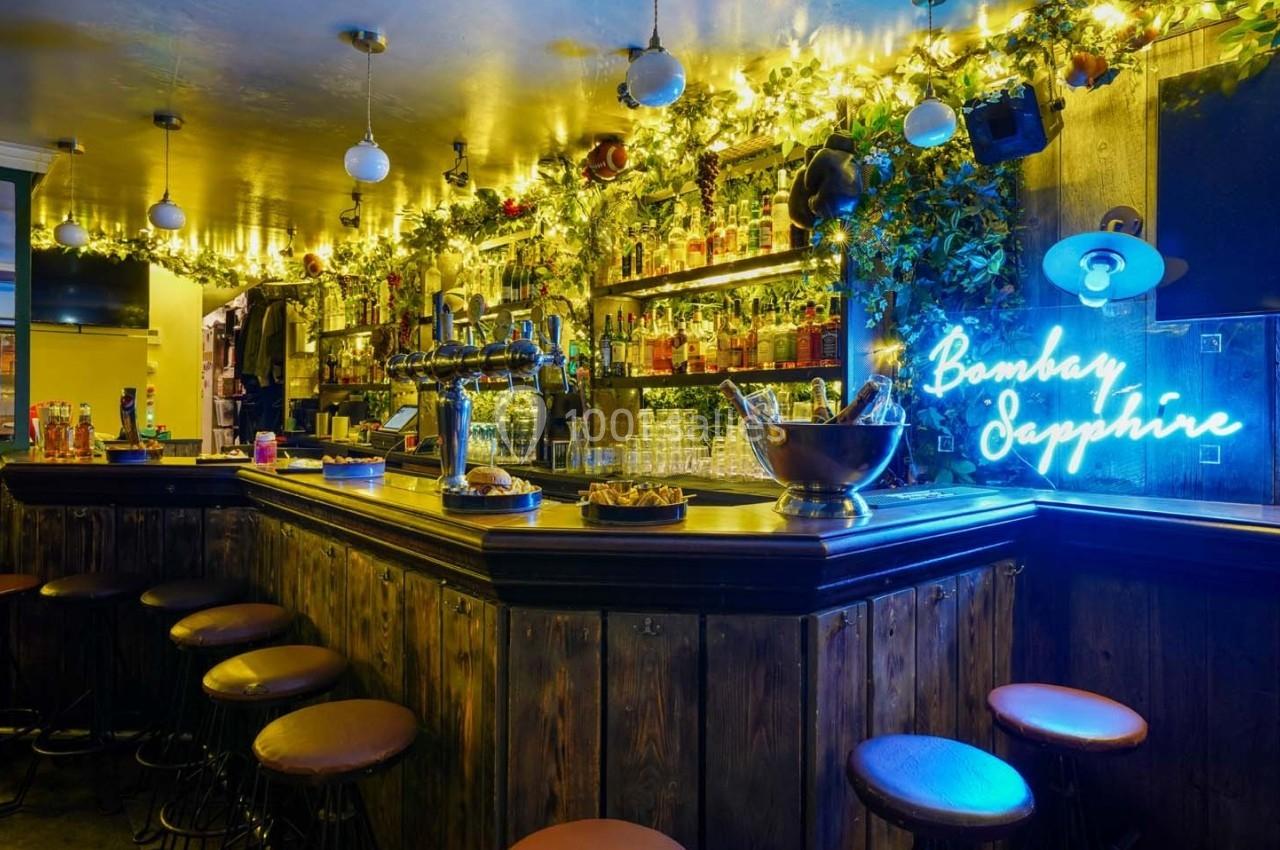 Bar en bois avec tabourets, étagères remplies de bouteilles, lumières décoratives et enseigne lumineuse bleue.