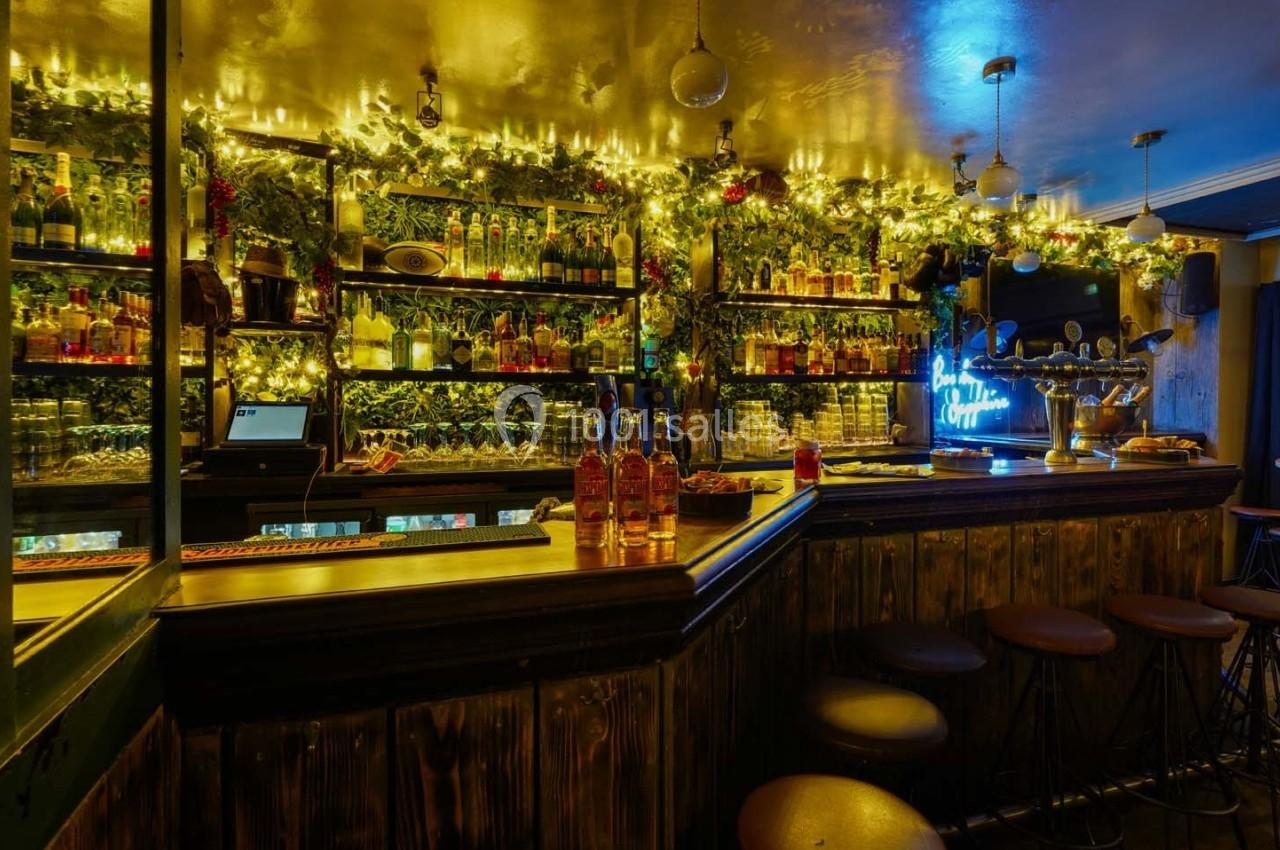 Bar avec comptoir en bois, étagères remplies de bouteilles, décorations lumineuses et ambiance chaleureuse.