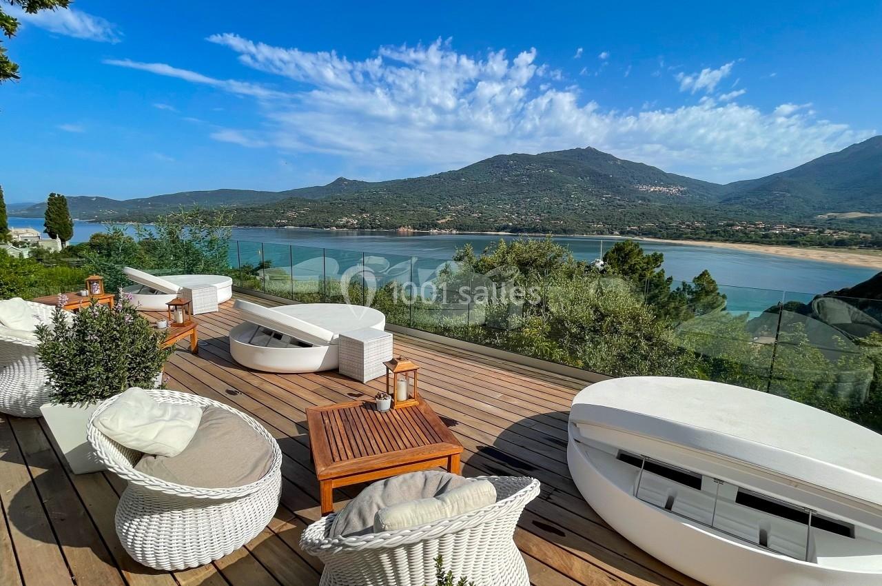 Terrasse en bois avec mobilier blanc, vue panoramique sur une baie entourée de montagnes sous un ciel dégagé.
