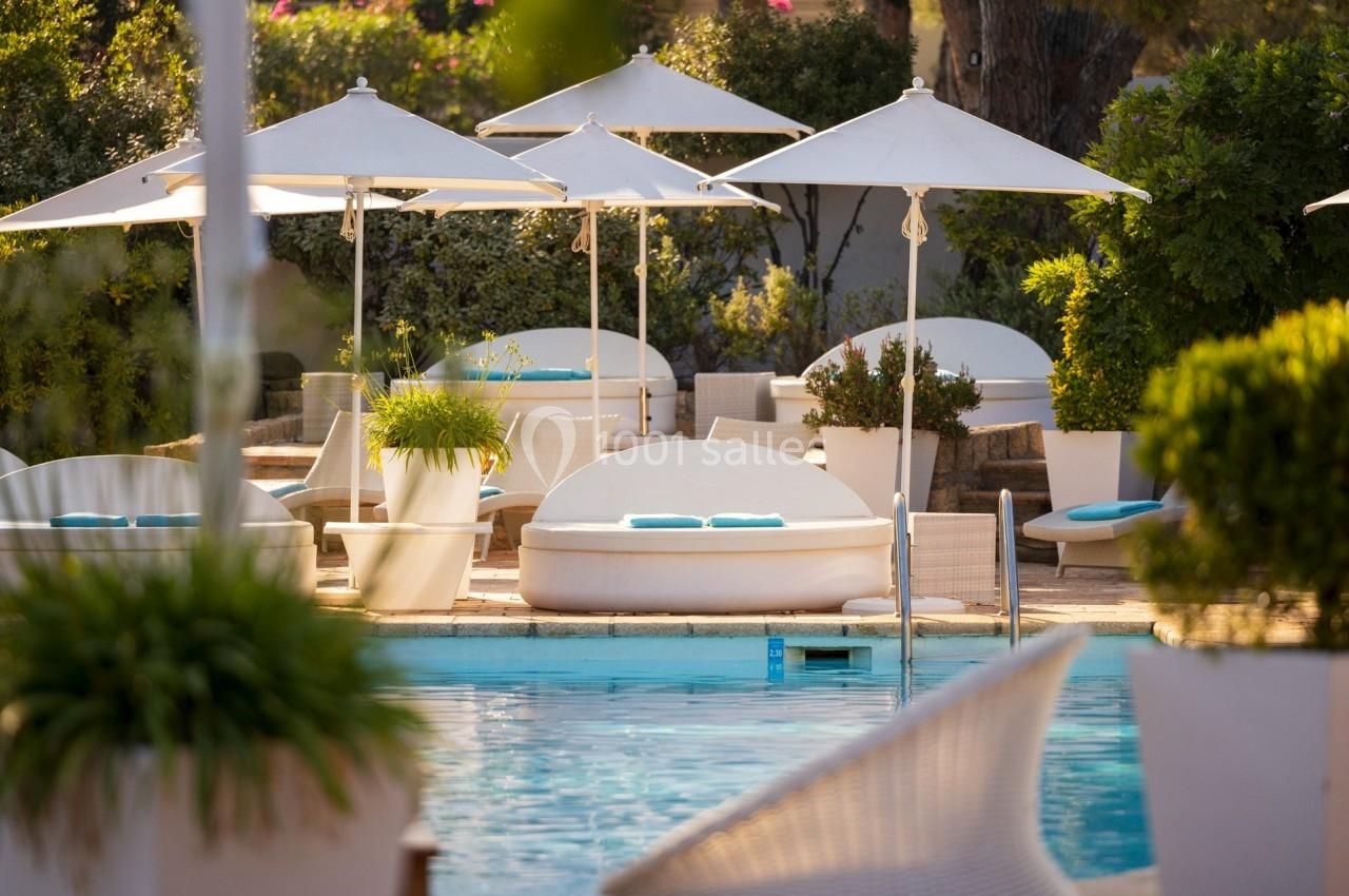 Piscine extérieure entourée de transats blancs, parasols et végétation dans un cadre paisible et ensoleillé.