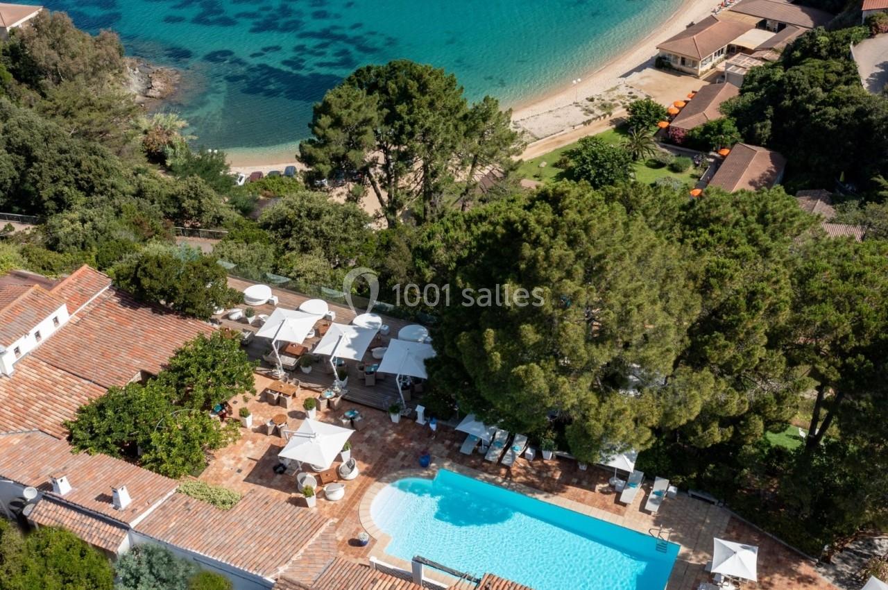 Vue aérienne d'une piscine entourée de terrasses et de végétation, près d'une plage bordée d'eau turquoise.