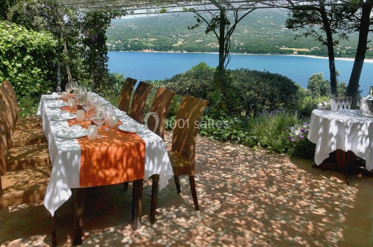 Table dressée avec nappe orange sous une pergola ombragée, vue sur un paysage de mer et de collines verdoyantes.